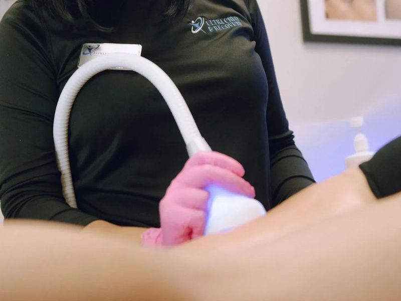 Woman in black shirt and pink gloves performing a body treatment with a device. Setting appears to be a medical spa.