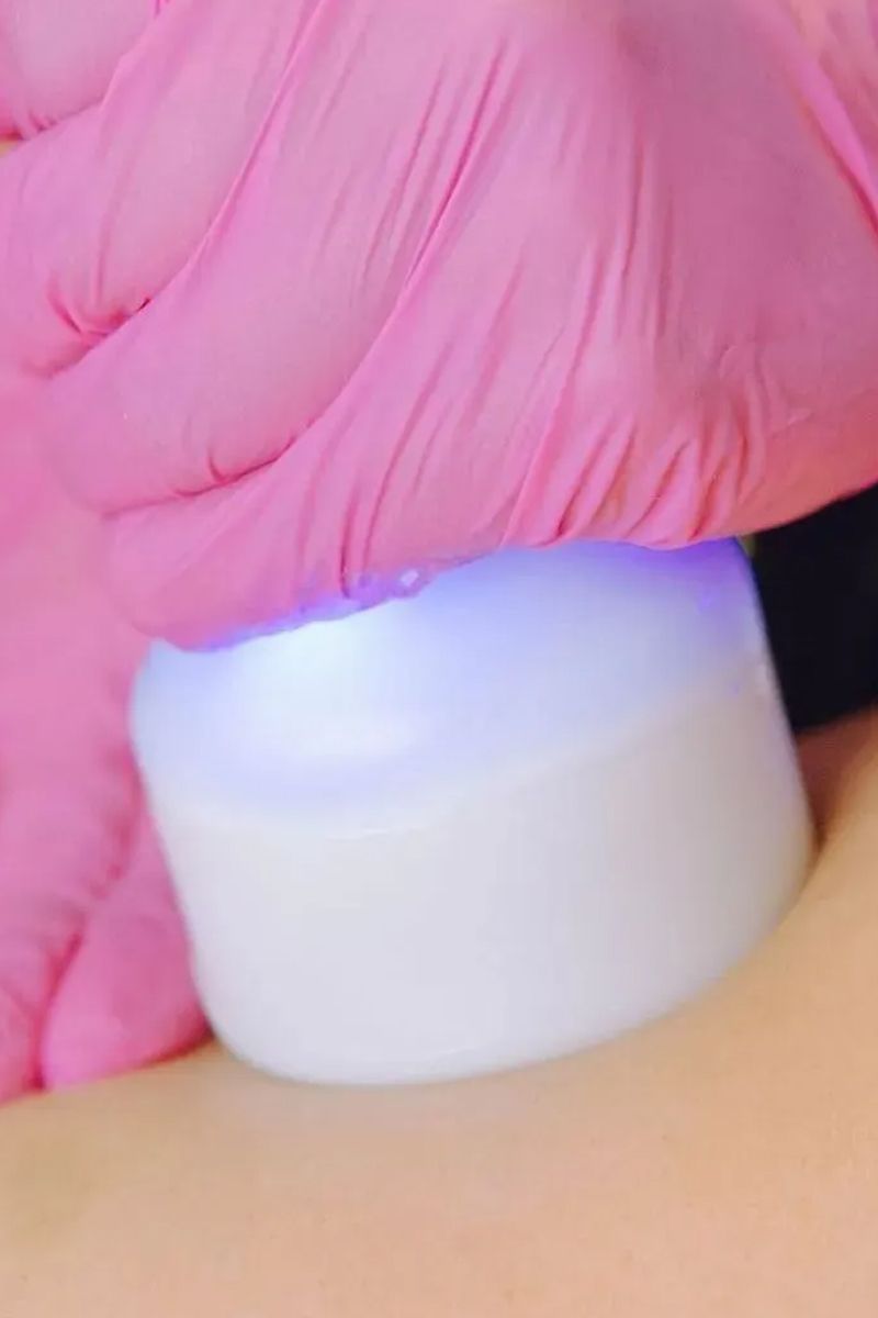 Pink-gloved hand holding a white device emitting a blue light, pressing it against skin.