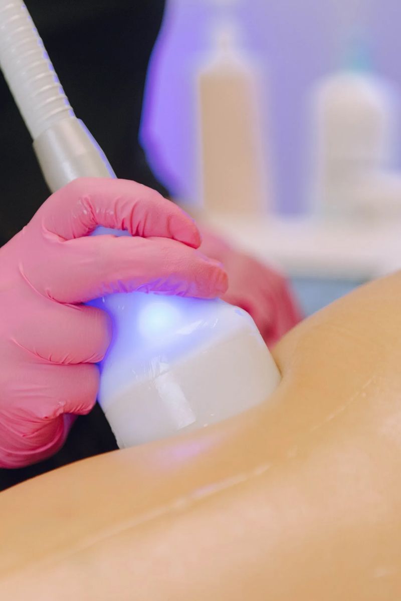 Person wearing pink gloves uses a white device on a leg, likely for a cosmetic procedure. Blue light is emitted from the device.