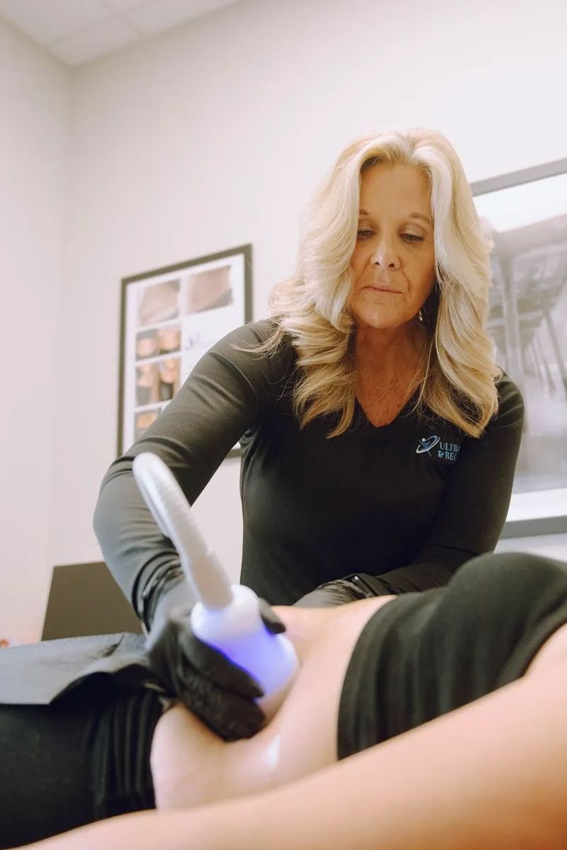 Person wearing pink gloves uses a white device on a leg, likely for a cosmetic procedure. Blue light is emitted from the device.