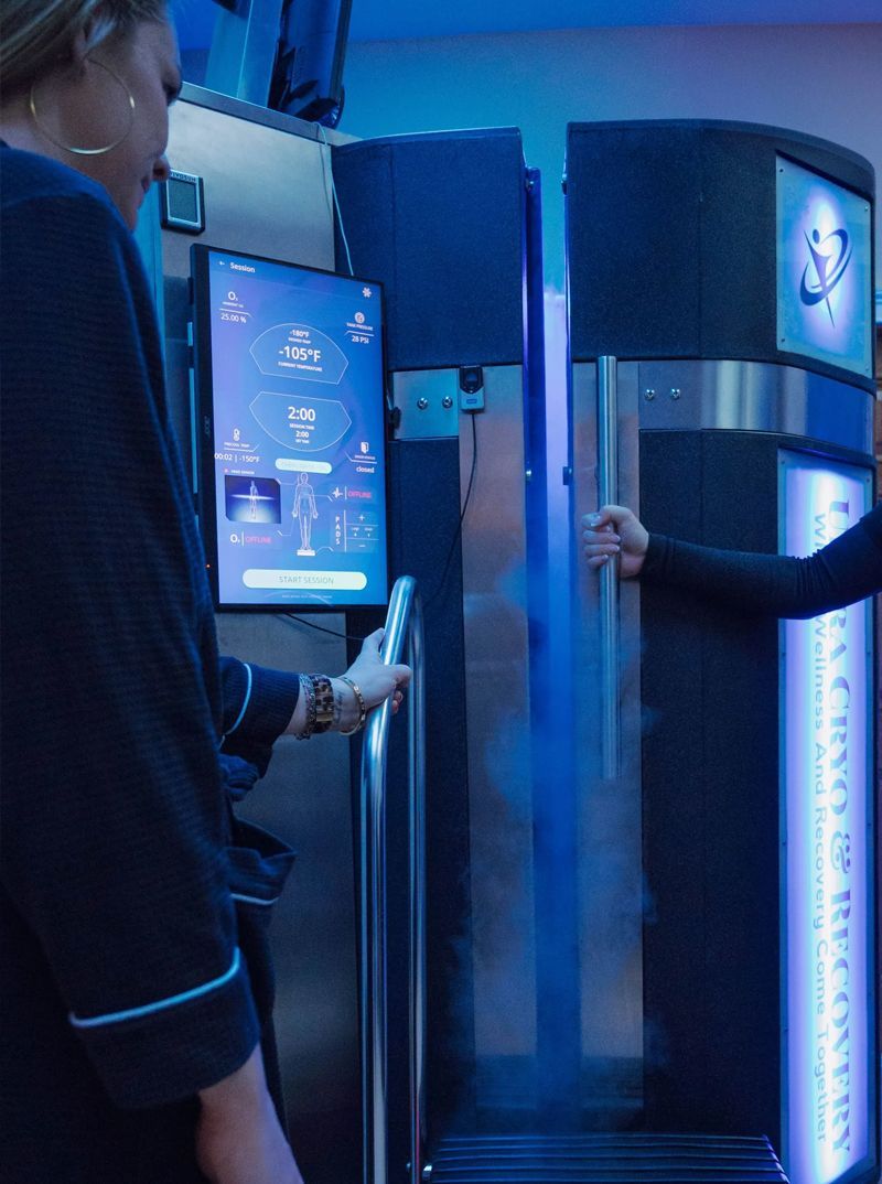 Woman in robe using a cryotherapy chamber. Cold fog billows out as the door opens.  A display screen is visible.