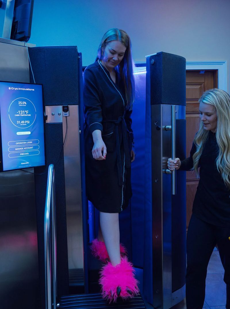 Woman in robe and fluffy pink slippers inside cryotherapy chamber, assisted by another woman in black.