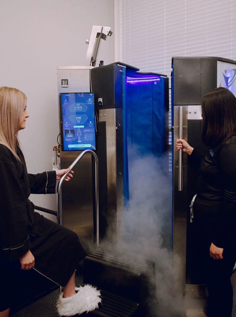 Two women stand near a cryotherapy machine, with one about to enter the chamber emitting cold vapor. The setting appears to be a medical spa.