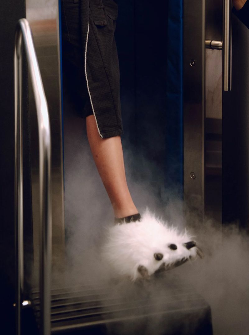 A woman in a black robe stands in a cryotherapy chamber, with vapor billowing out.