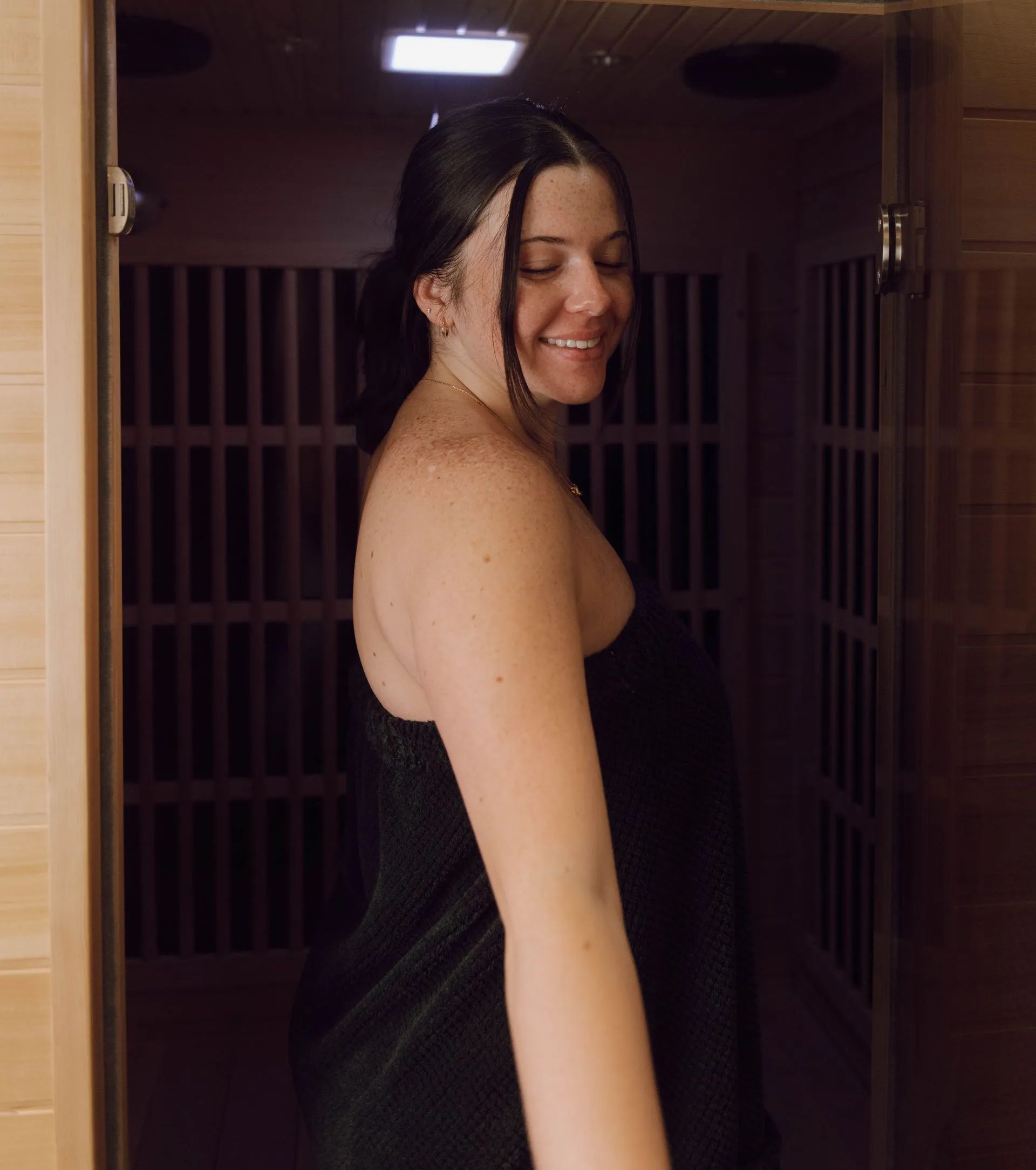 Woman wrapped in a black towel smiles inside a wooden sauna. Her dark hair is pulled back.