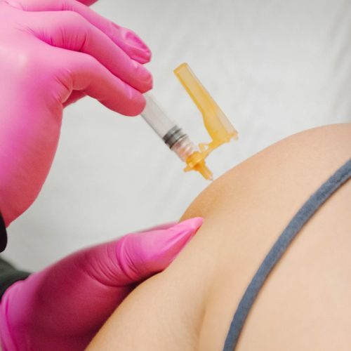 A person in pink gloves giving an injection into the upper arm of another person wearing a gray tank top.