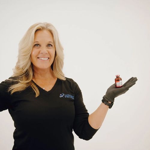 Woman with blonde hair, wearing a black top and glove, smiling and holding up a small brown bottle against a white background.