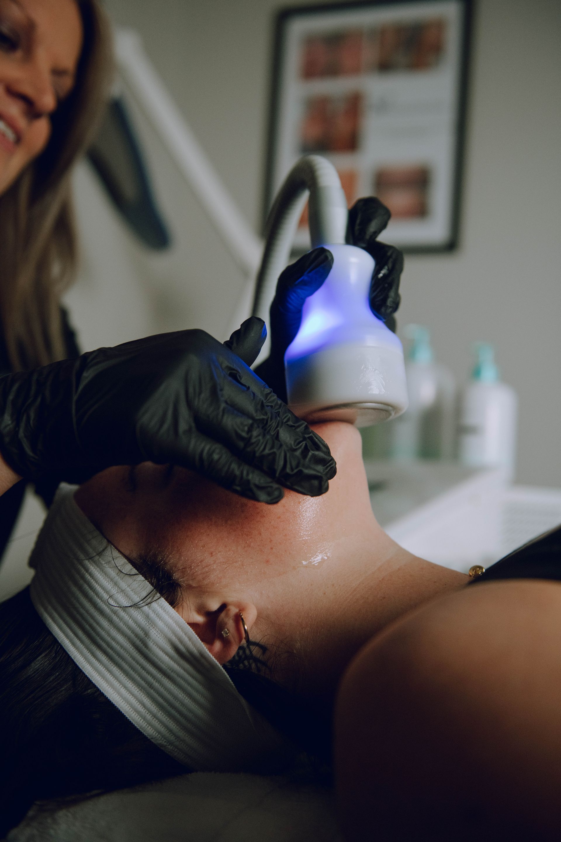 Person receiving facial treatment; technician holding device to the person's neck. Dark gloves, white light, indoor setting.