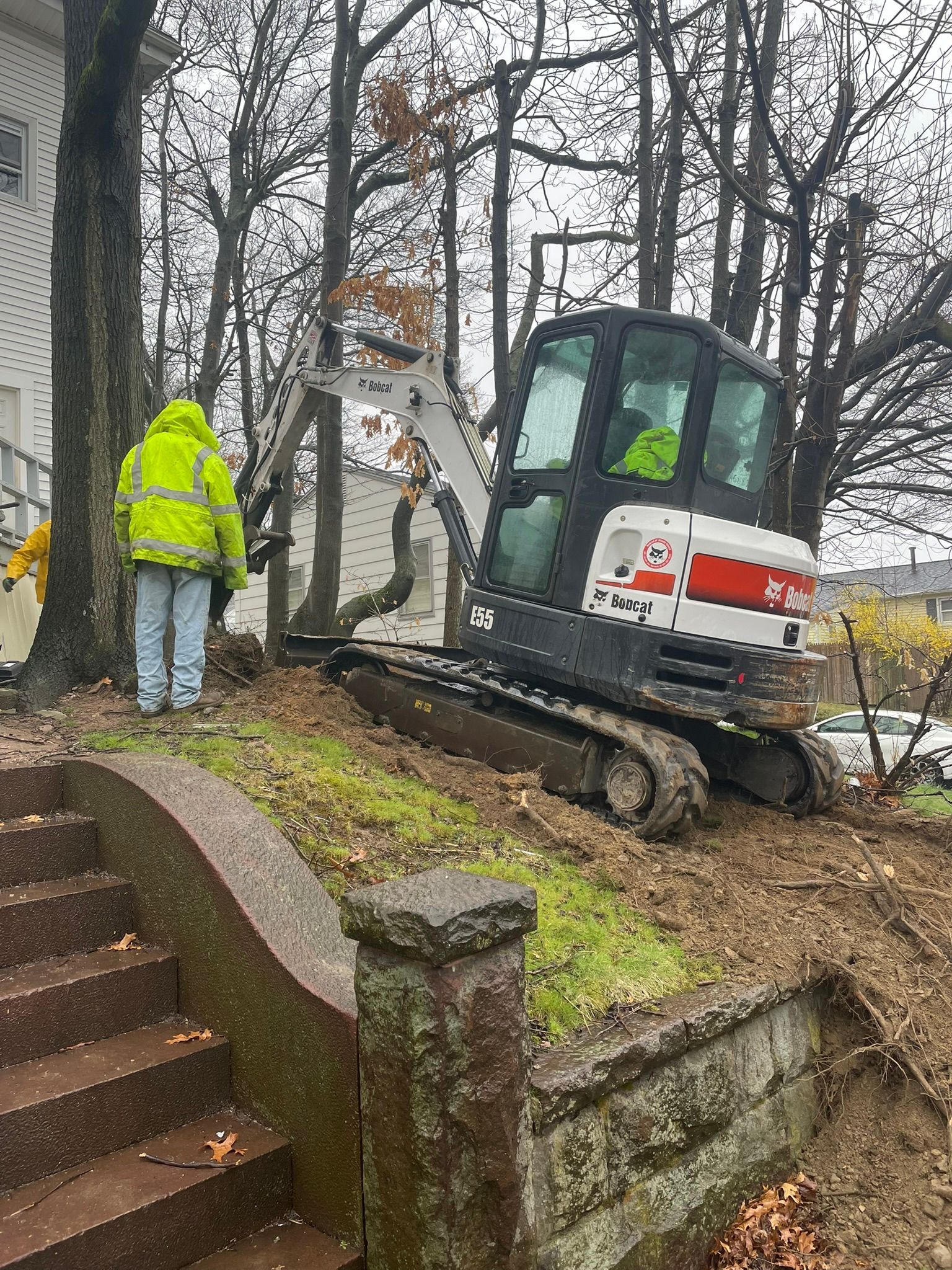A man in a yellow jacket is standing next to a small excavator.