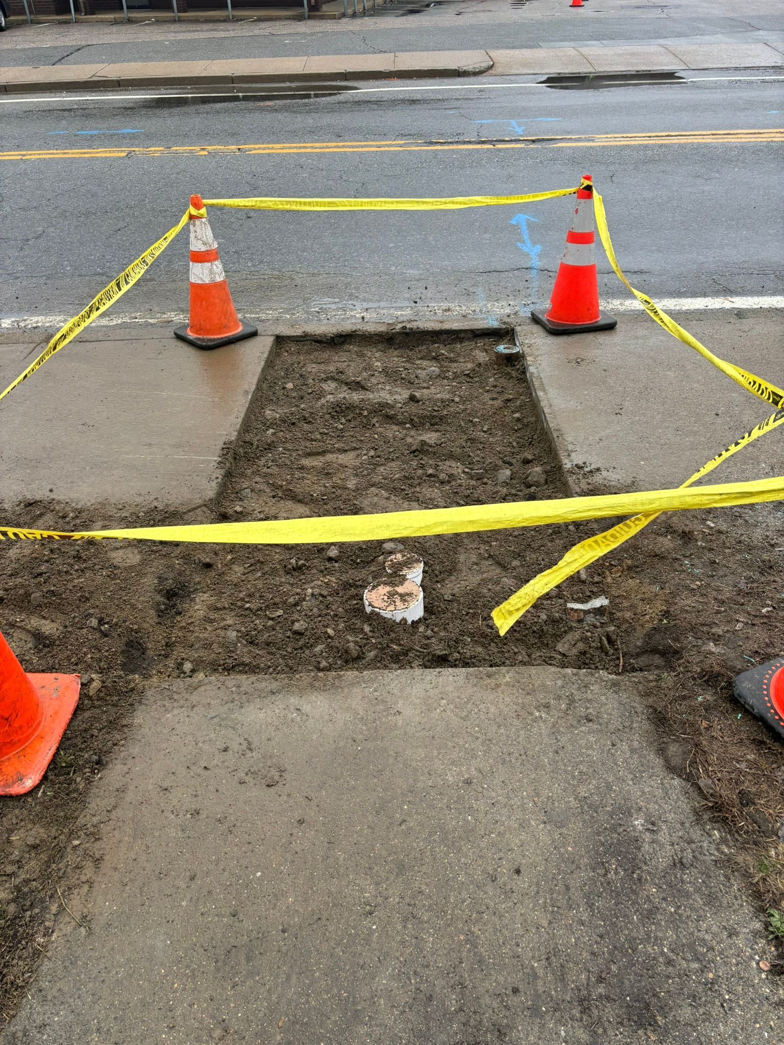 A hole in the sidewalk is surrounded by orange traffic cones and yellow tape.