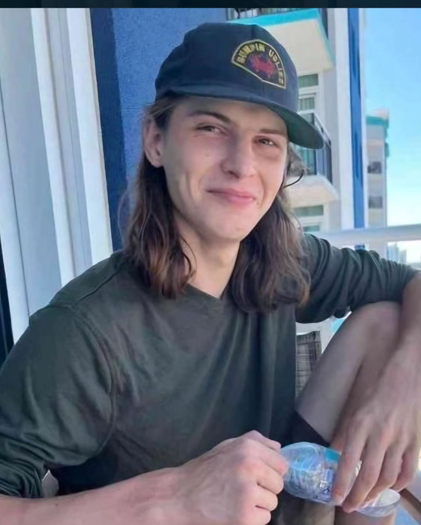 A young man with long hair is wearing a hat and holding a bottle of water.