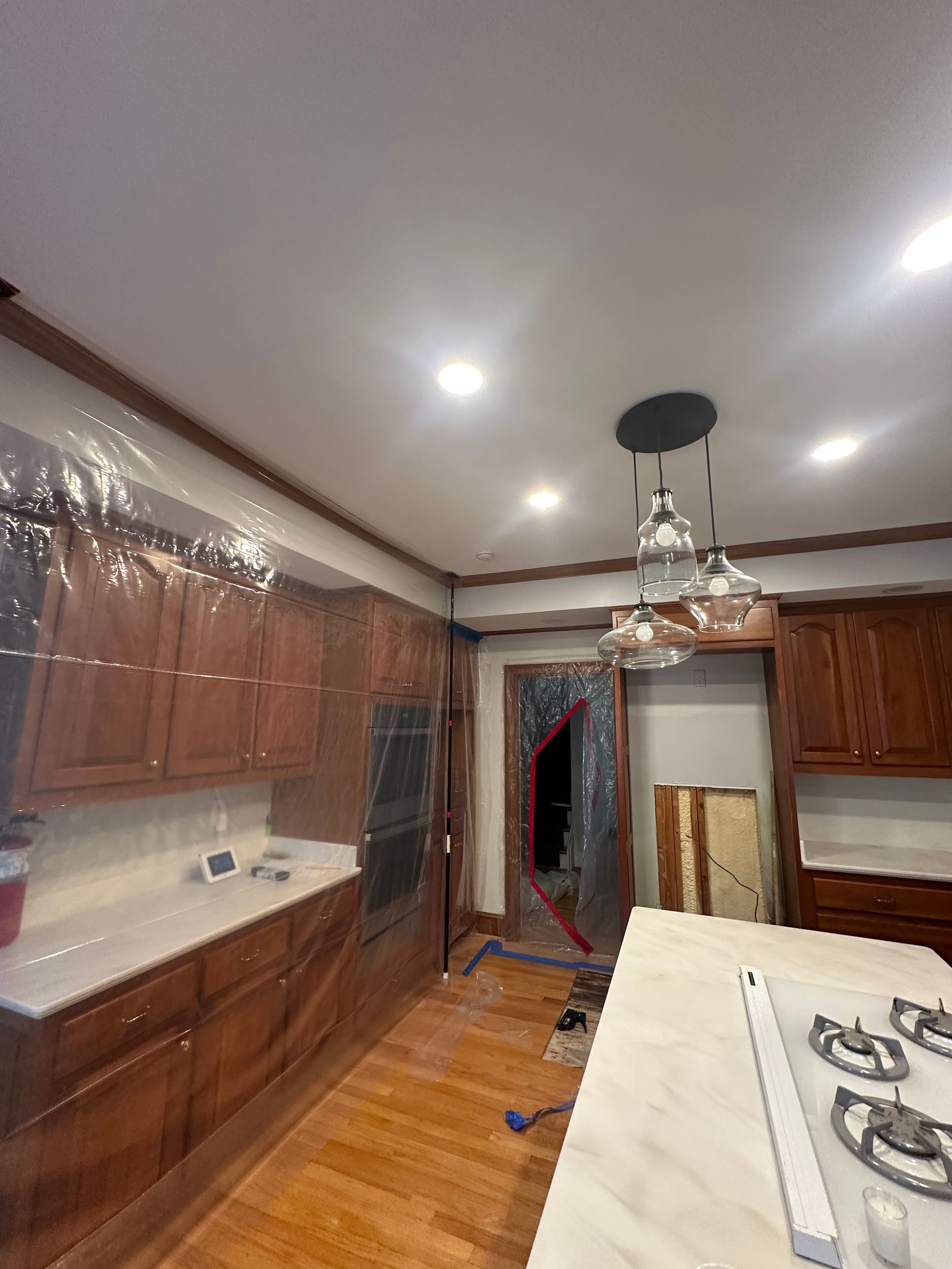 Kitchen under renovation, covered with plastic sheeting. Brown cabinets, wooden floor, and a stove are visible.