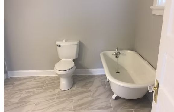 Bathroom with a white toilet and a claw-foot bathtub on gray tiled floor, with gray walls and white trim.
