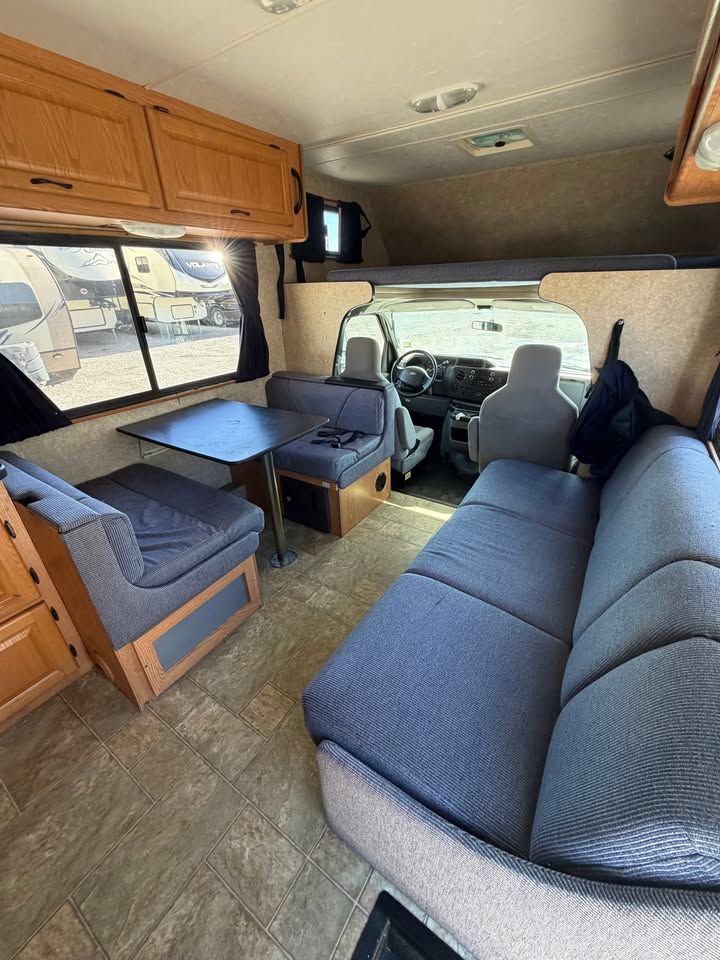 Interior of an RV with blue seating, table, wooden cabinets, and a view of the driver's area.