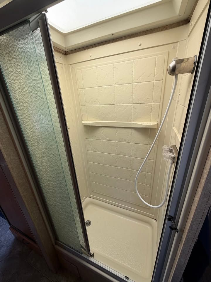 RV shower stall with a frosted glass door, shelf, and showerhead. Beige walls and white basin.