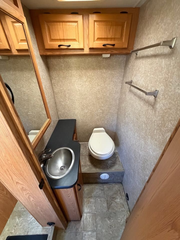 Small RV bathroom with toilet, sink, mirror, and cabinets. Beige walls, light wood cabinets, and gray floor.