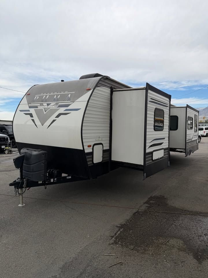 White and black travel trailer with open slide-out in a paved lot under a cloudy sky.