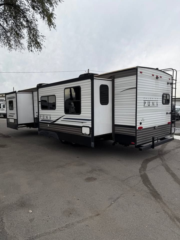 RV trailer with two slide-outs, white and gray siding, parked on asphalt, cloudy sky.