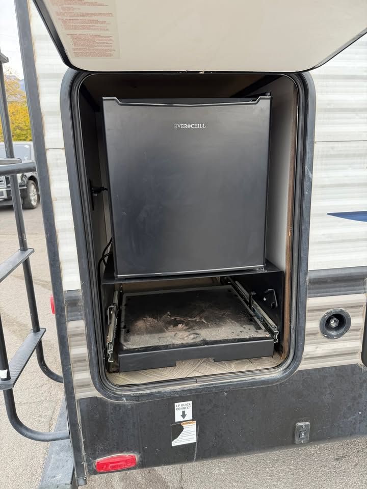 RV exterior storage compartment with a black mini-fridge and a pull-out grill on a slide.