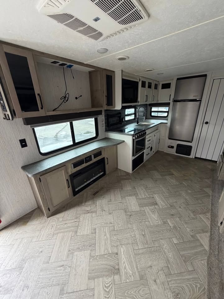 Interior of a camper with kitchen, fireplace, cabinets, and appliances; light wood flooring.