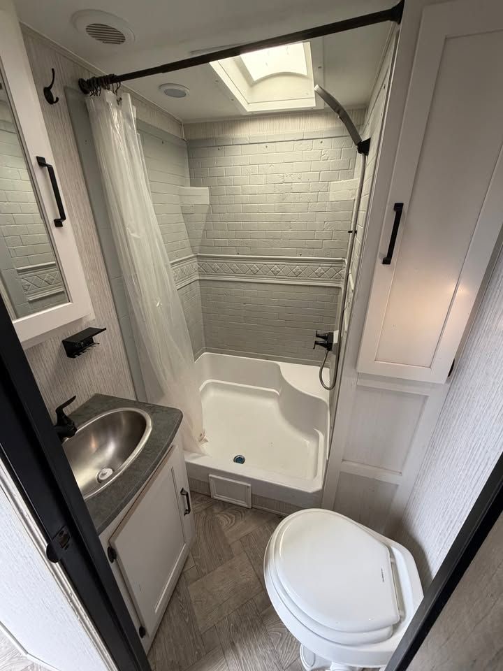 RV bathroom with a shower, toilet, sink, and storage; gray and white color scheme.