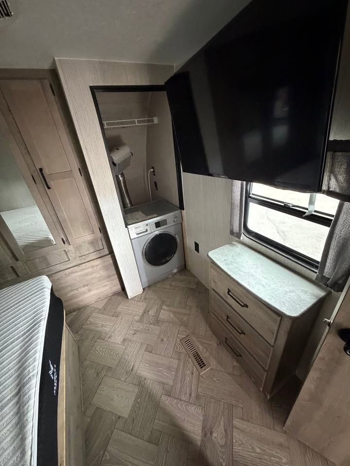 RV interior with washer, TV, and dresser. Neutral tones, herringbone floor, window.