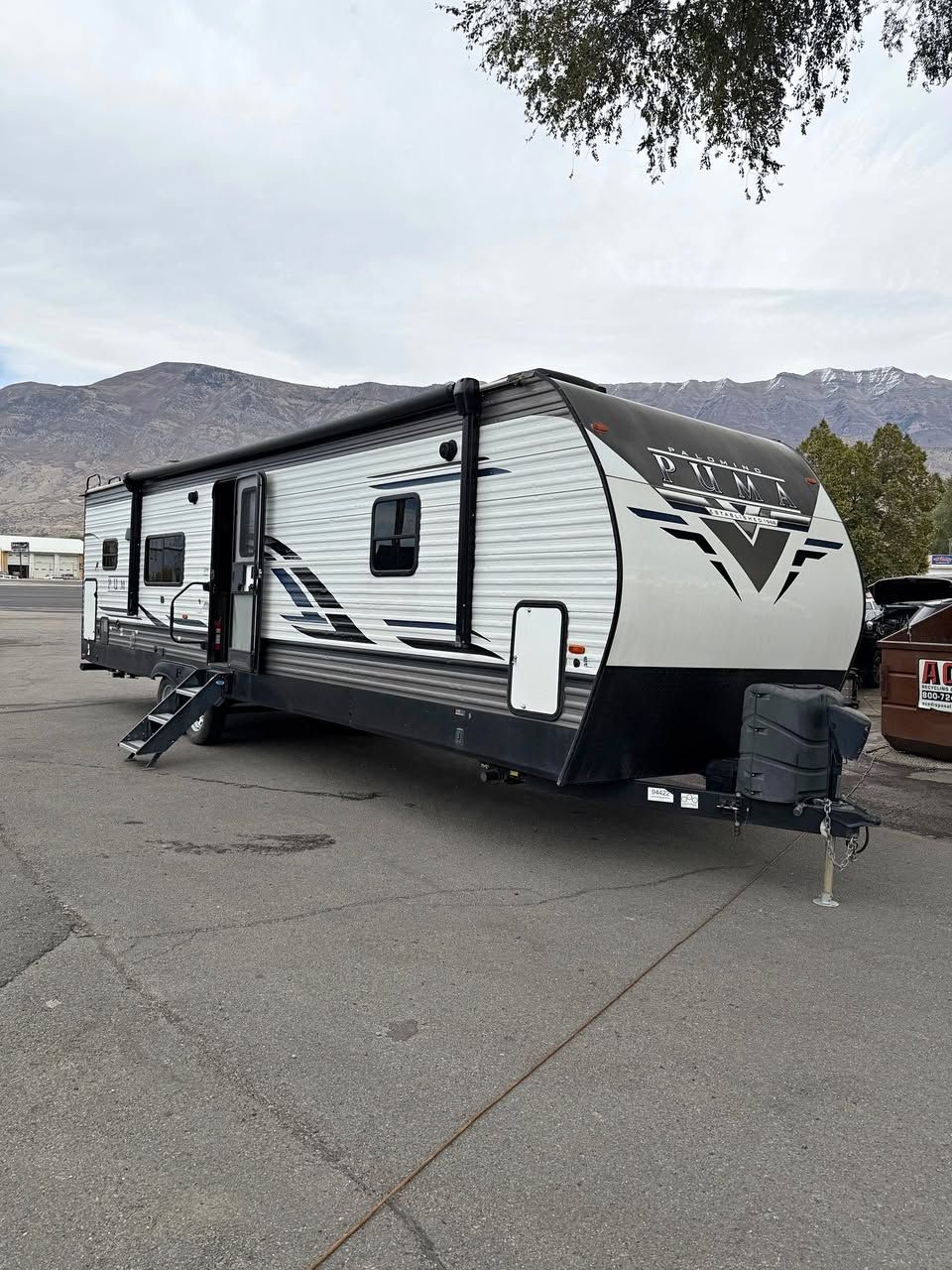 Gray and white RV trailer parked on pavement, mountains in the background. Black awning extended.