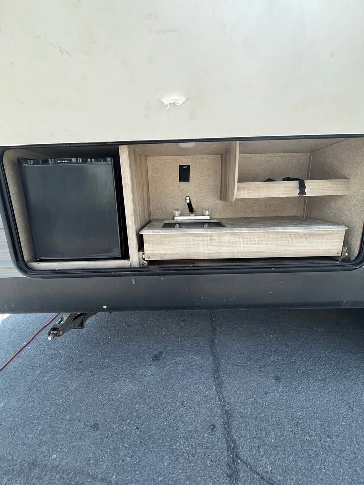 RV outdoor kitchen: small refrigerator, sink, counter, shelves, and electrical outlet.