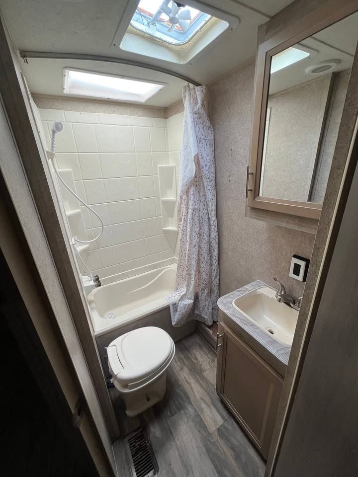 Small RV bathroom with toilet, tub, sink, and mirror. Beige and white interior, with a vent in the ceiling.