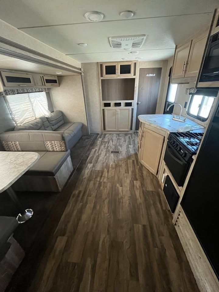 Interior of a travel trailer with kitchen, dining area, and sofa. Wooden floors and light-colored cabinets.