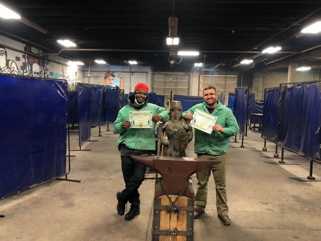 Two people are standing next to an anvil in a factory holding certificates.