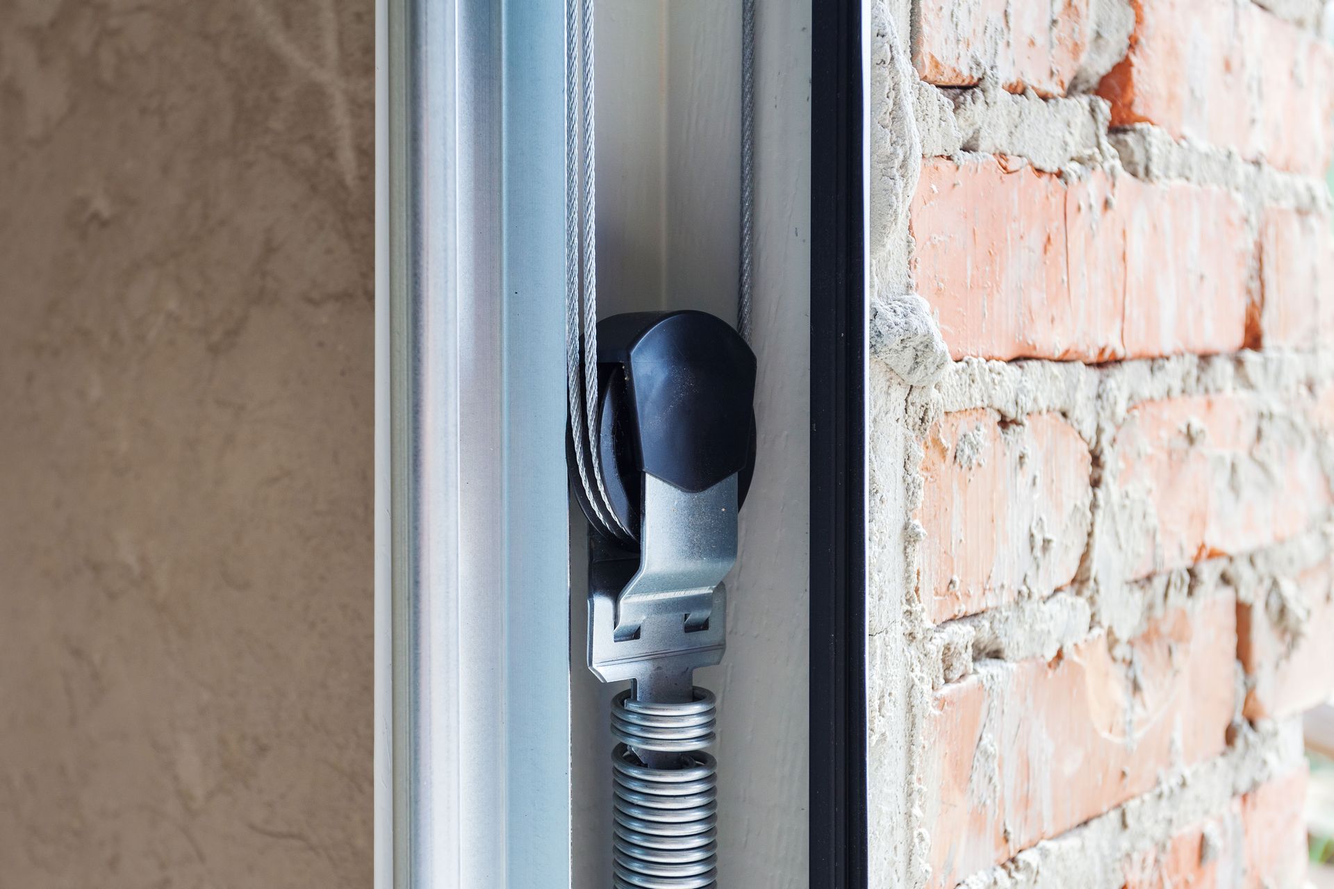 Close-up of a window blind mechanism with a spring, pulley, and cable, set against a brick wall.