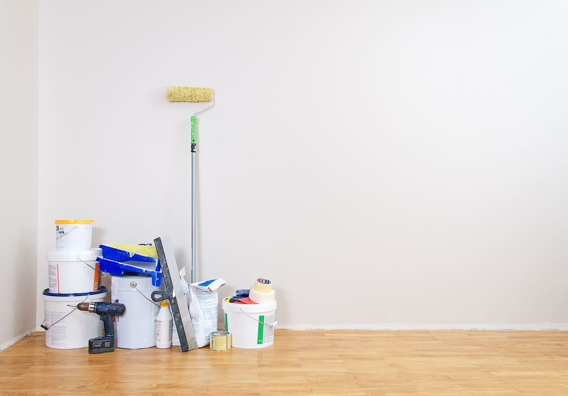 Painting supplies (paint cans, roller, brushes, drill) rest against a white wall in a room with a wood floor.