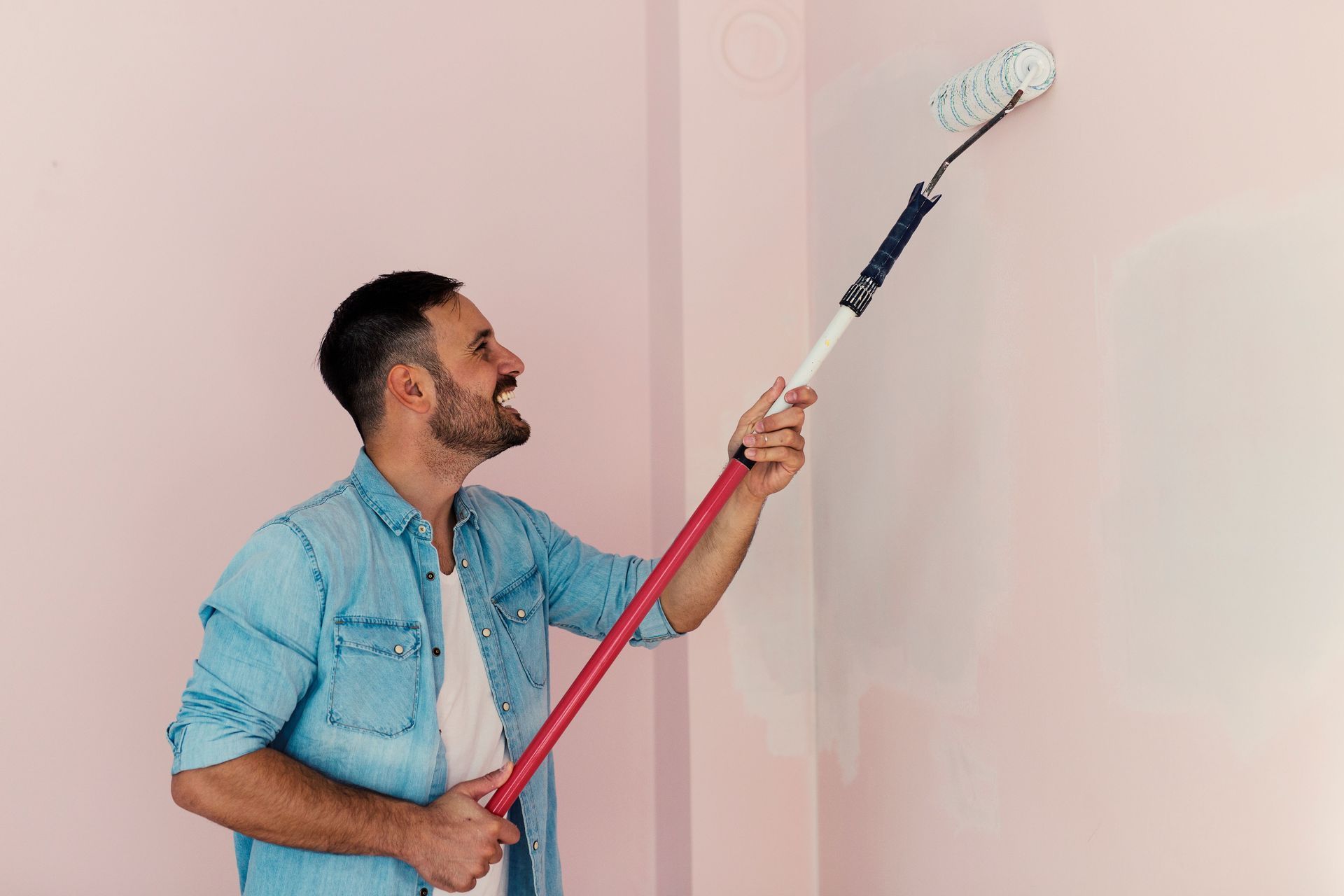 Man painting a pink wall with a roller. He has a beard and is looking up.