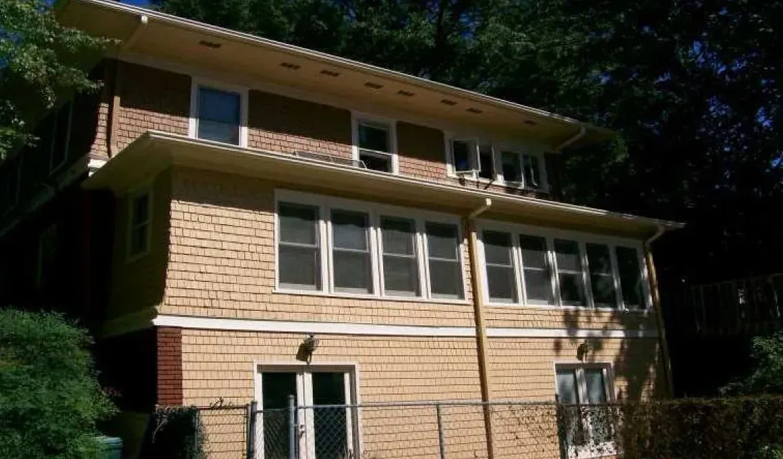 Two-story light brown house with multiple windows and a small yard, partly obscured by trees.