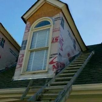 Dormer window on roof under construction, covered in protective wrap, with a ladder leaned against the roof.