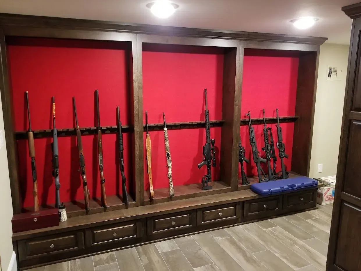 Gun rack with rifles displayed against a red background, with drawers and bench below.
