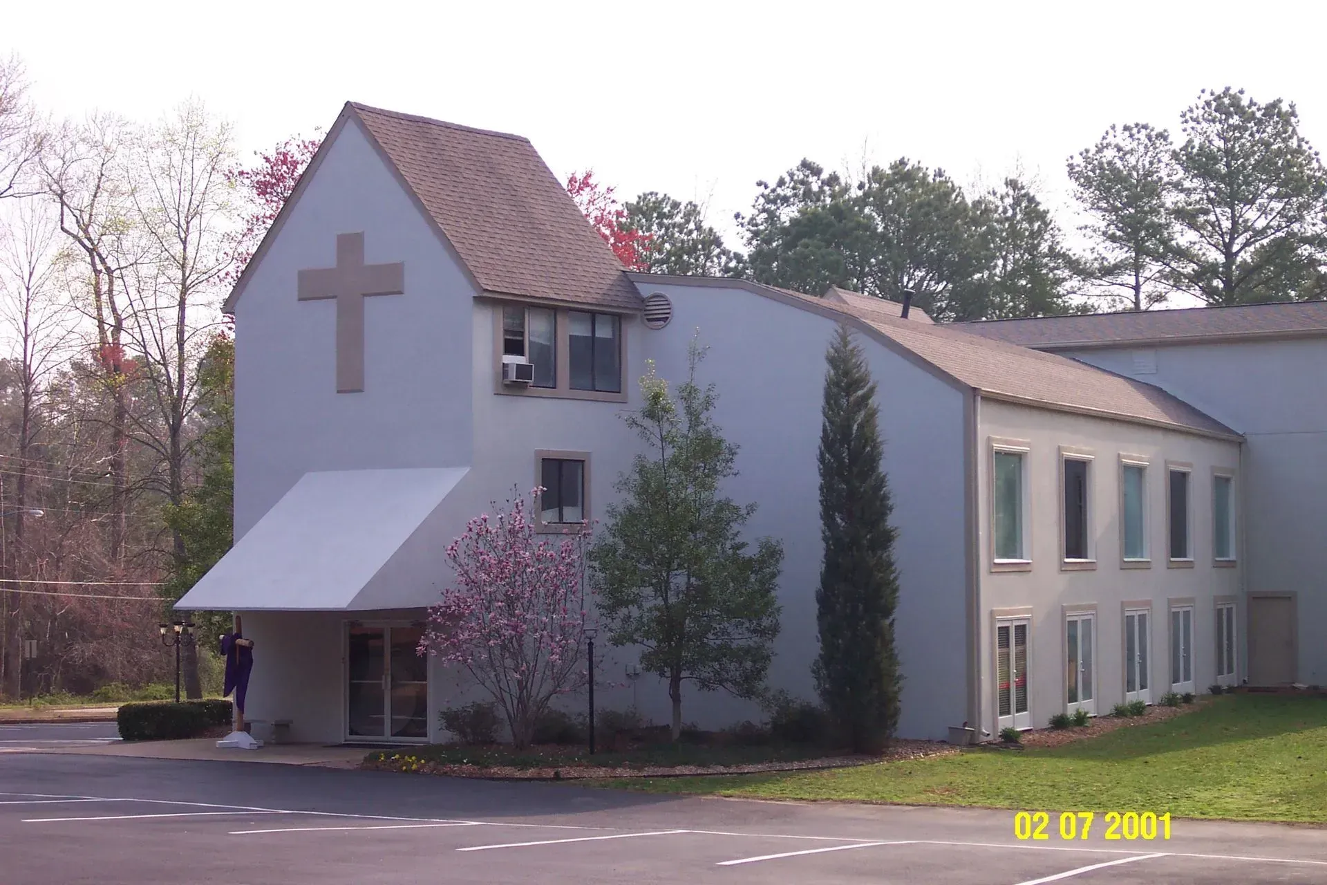 Church building with a cross, white exterior, trees, and blue sky.