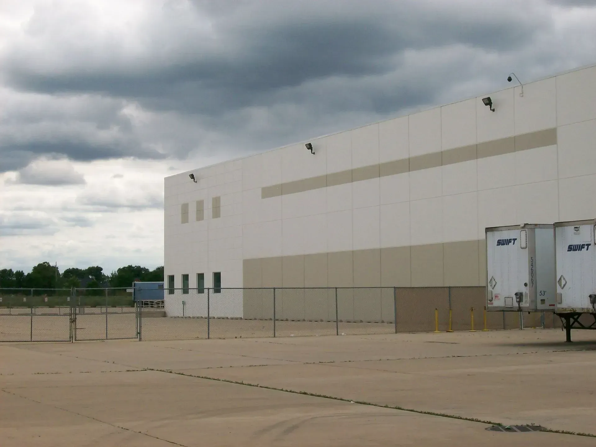 Large, white industrial building with a cloudy sky. Two semi-trailer trucks are parked on the side.