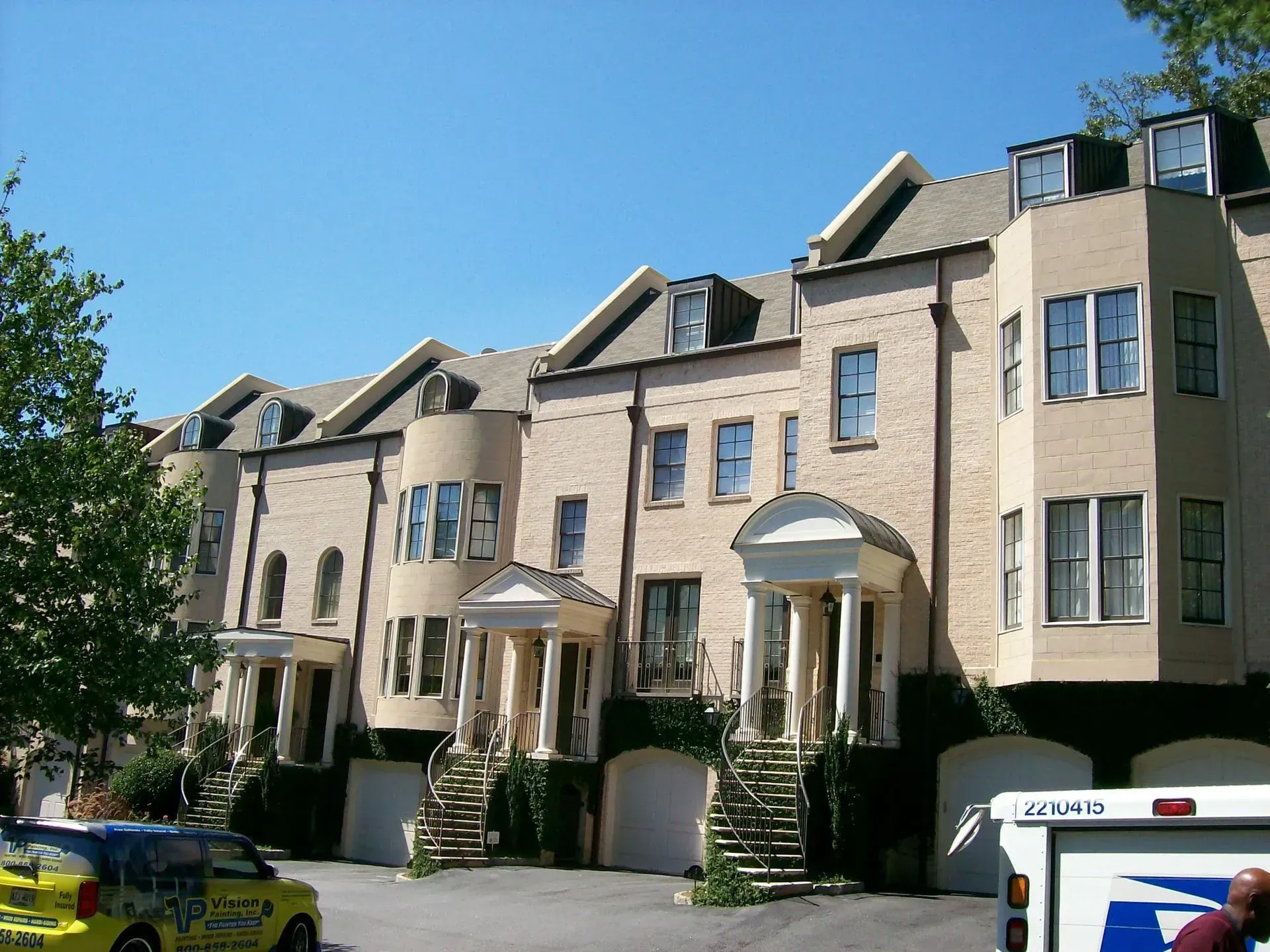 Row of beige townhouses with arched entryways, windows, and dark roof on a sunny day.