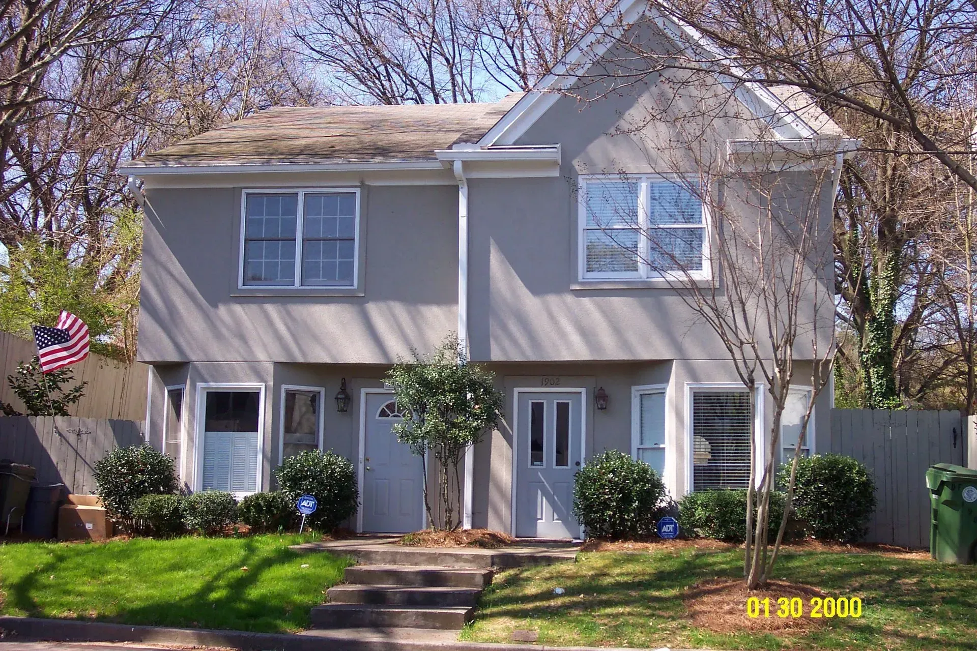 Two-story duplex with gray stucco siding, two front doors, windows, and an American flag on a sunny day.