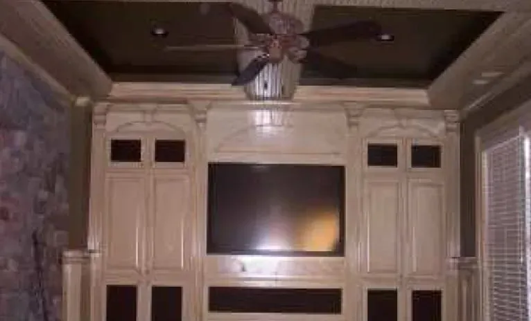 Built-in entertainment center with a large TV and cabinetry, beige and dark brown tones, ceiling fan.