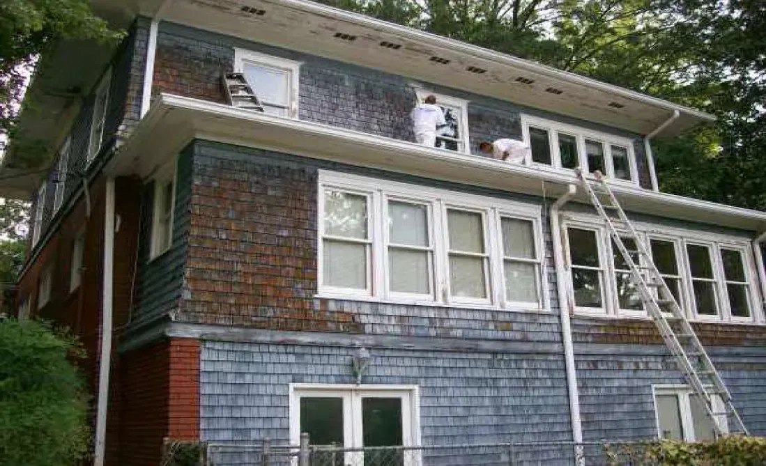 Two painters painting a two-story house with blue siding and white trim. Ladder visible.