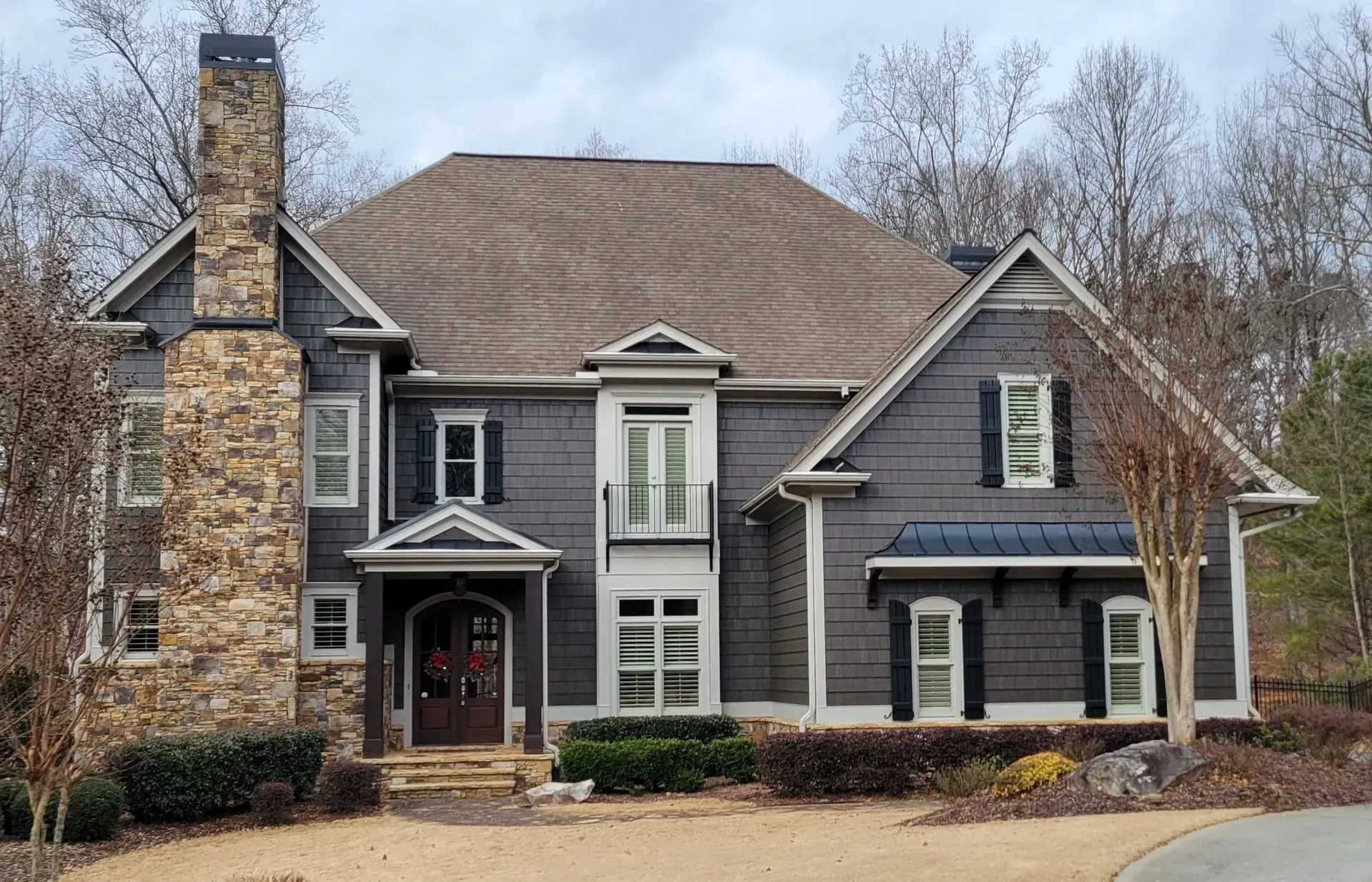 Two-story gray house with brown roof and stone chimney, set in a yard with trees.