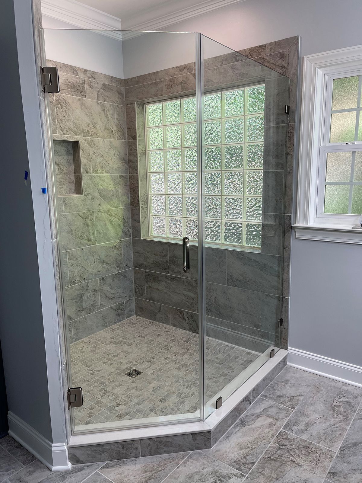 A modern walk-in shower with a glass door, grey stone tile walls, a pebble floor, and a glass block window.