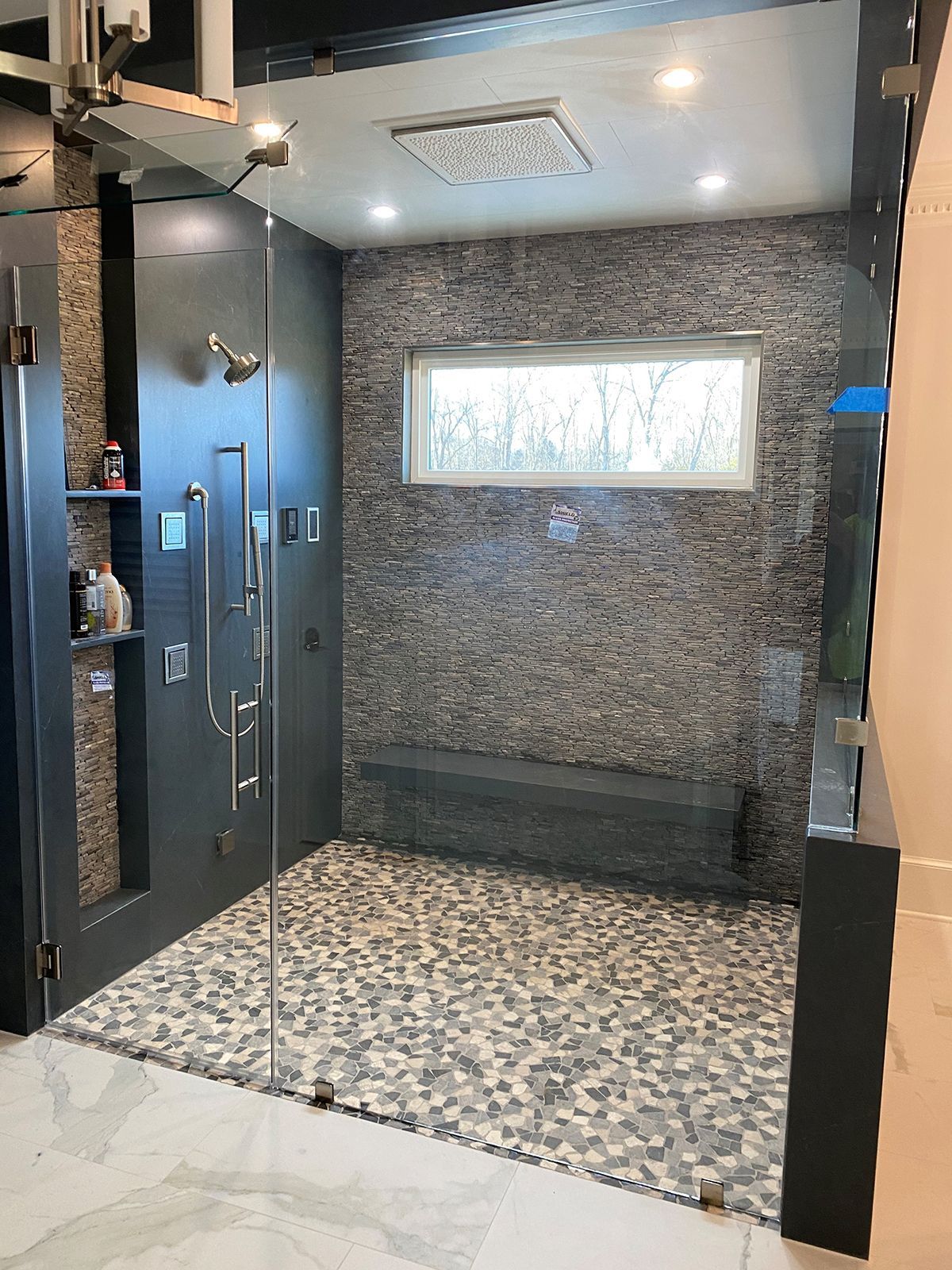 A modern walk-in shower with stone tile walls, a built-in bench, a pebble floor, glass doors, and a rain shower head.