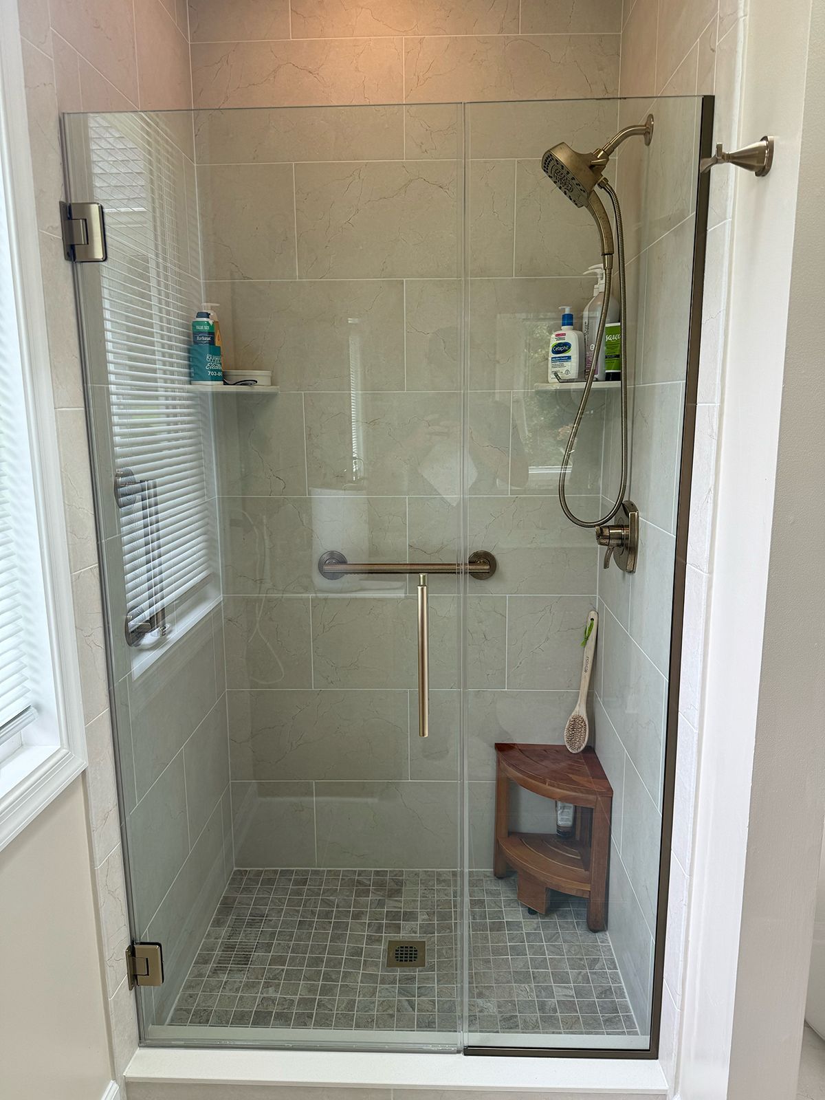 A walk-in shower with a glass door, light-colored tile walls, bronze fixtures, a grab bar, and a wooden corner stool.