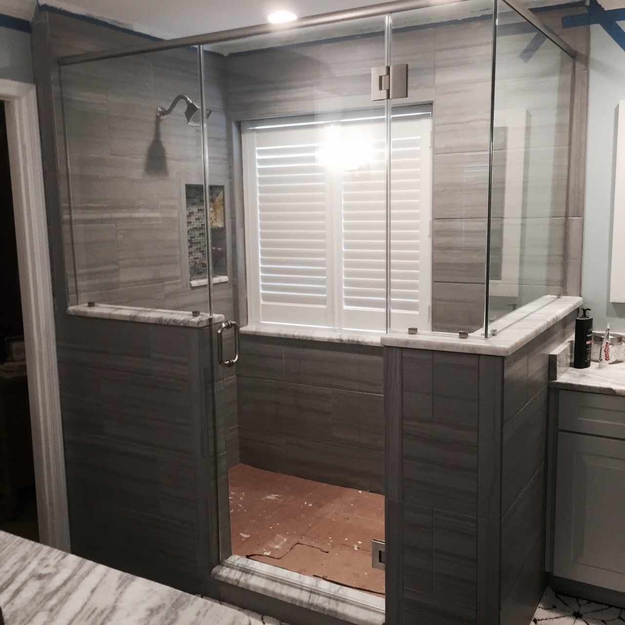Gray tiled shower with glass walls and door, white blinds behind.