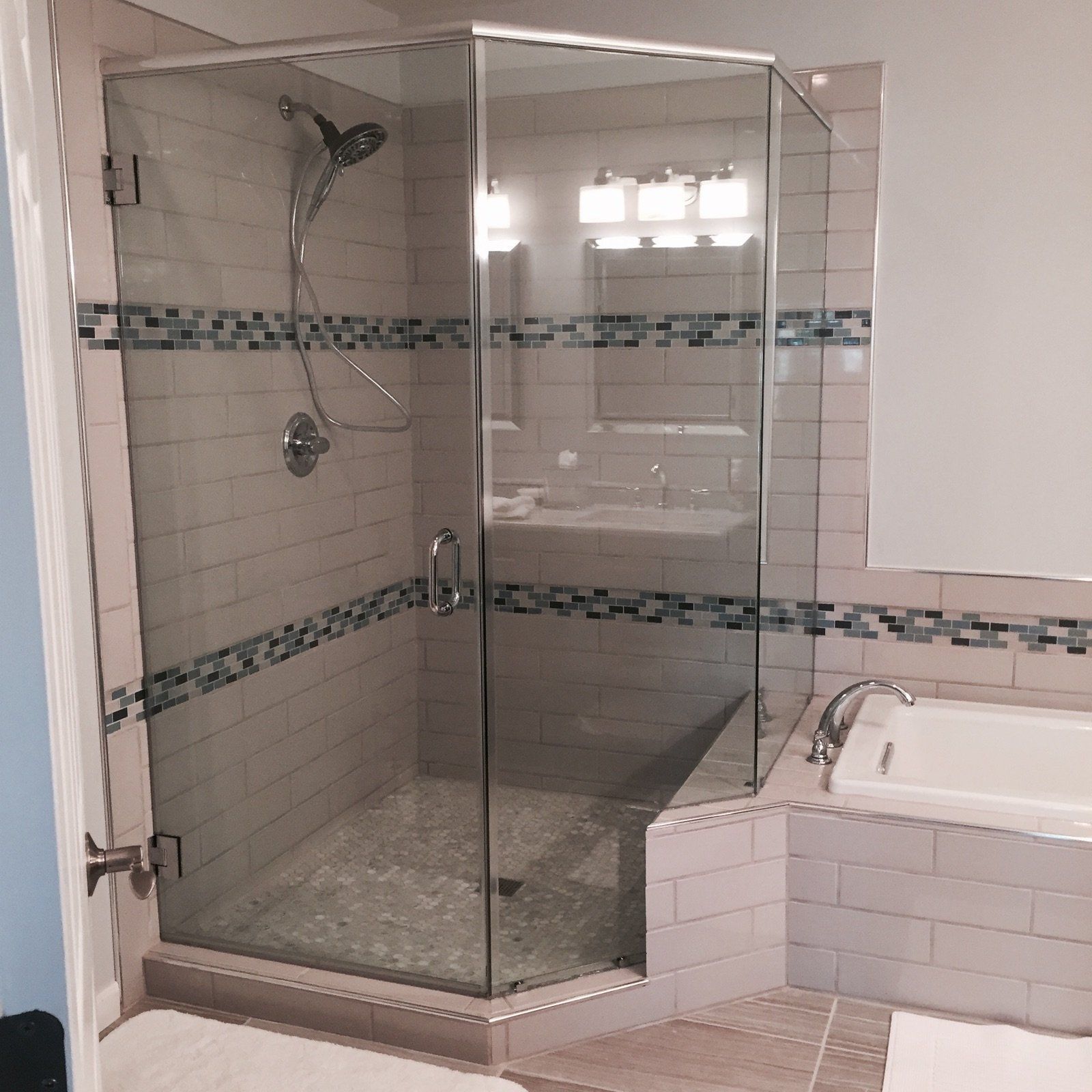 Glass shower enclosure in a modern bathroom with tiled walls and a small tub.