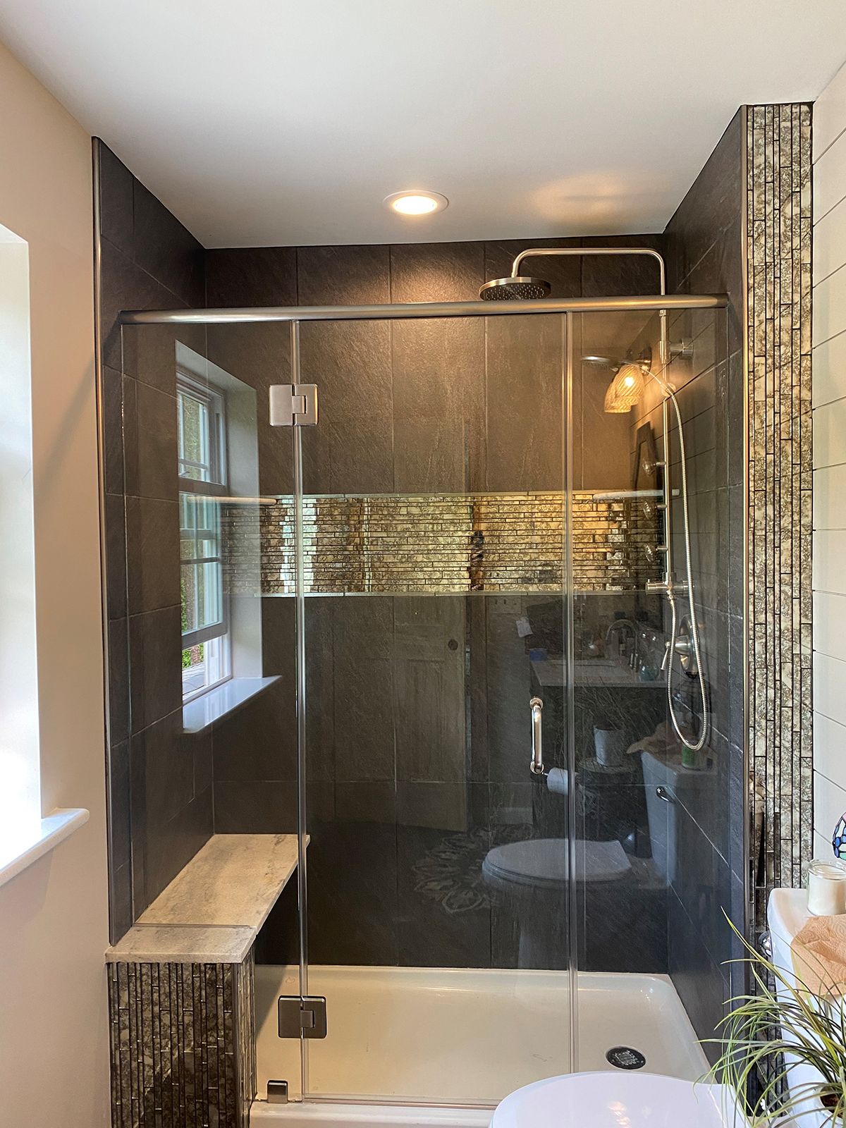 A modern shower with a glass sliding door, dark tiled walls with a gold accent stripe, and a built-in stone bench.